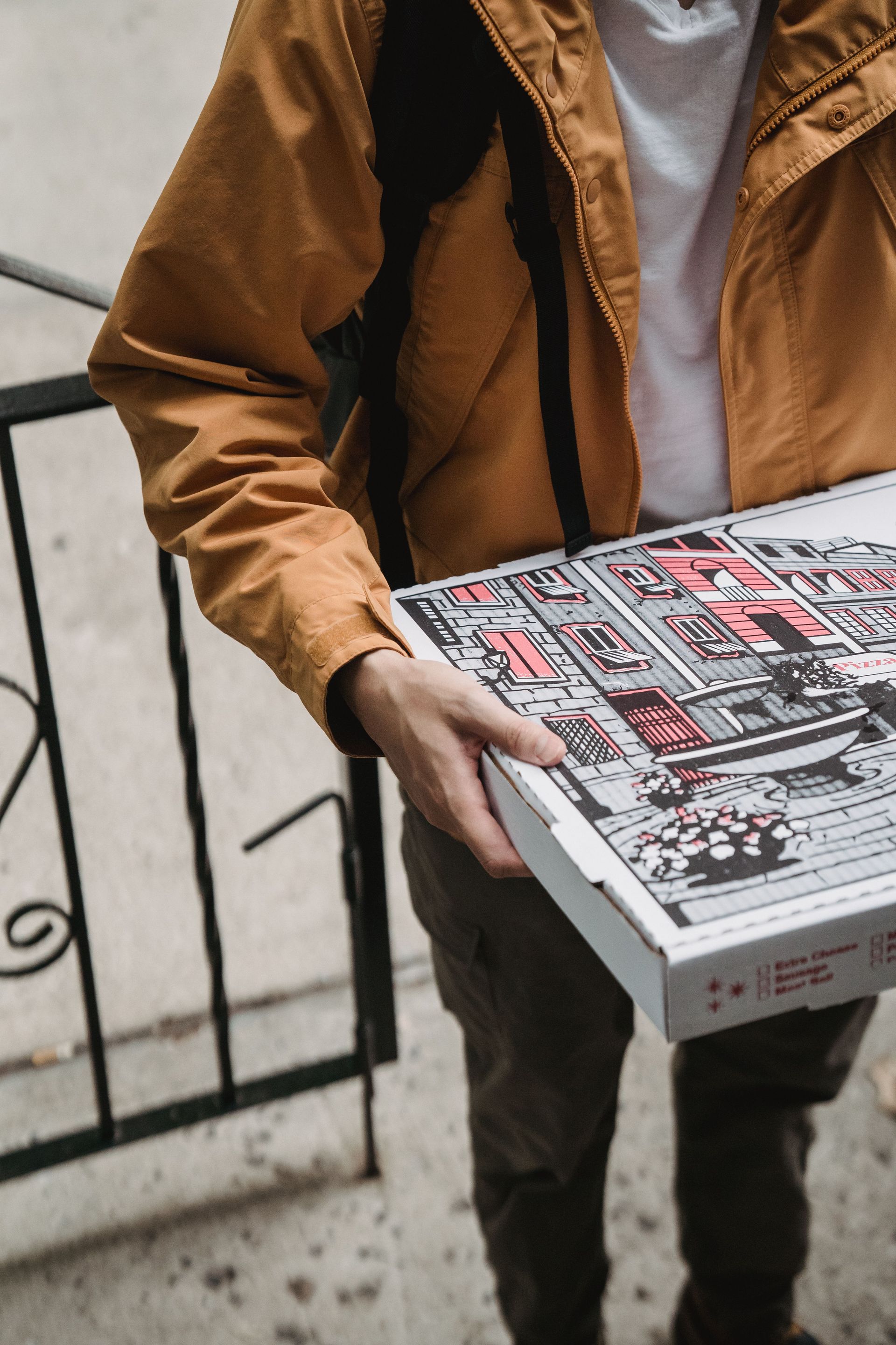 Close-up of a delivery person with a pizza box at the doorstep, wearing a brown jacket.